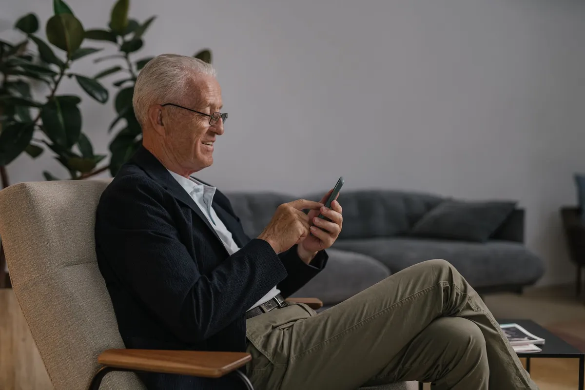 An older adult sitting in a living room chair with a phone within reach on a side table
