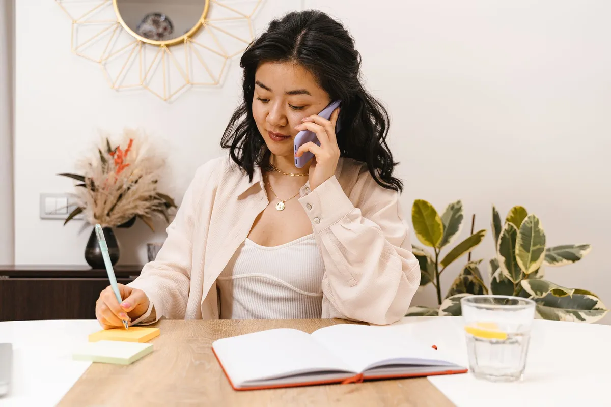 Person on a phone call at a kitchen table taking notes on a notepad