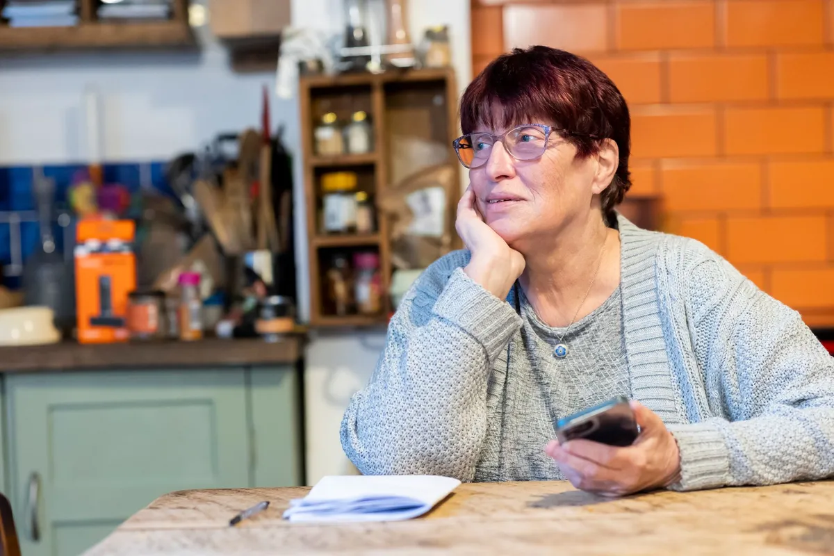 An older woman wearing glasses and a gray sweater sits at a kitchen table resting her chin on her hand while holding a smartphone, with papers and a pen in front of her