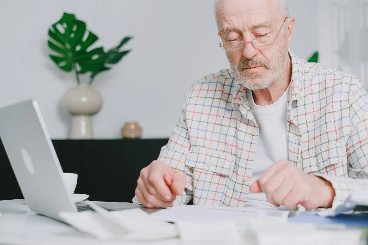 An older man wearing glasses looks down at paperwork on a table while sitting in front of an open laptop