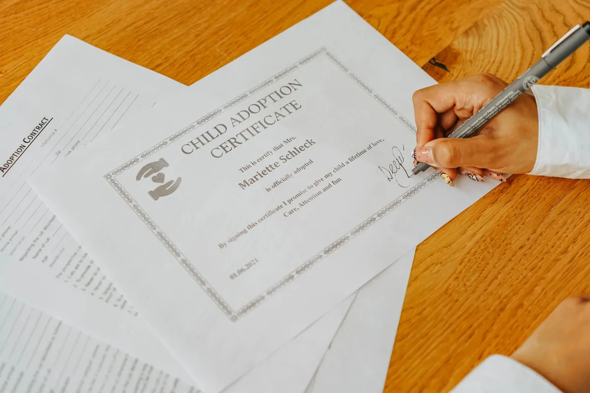 A person signs a printed Child Adoption Certificate for Mrs. Mariette Schleck with a pen, placed atop an adoption contract on a wooden table