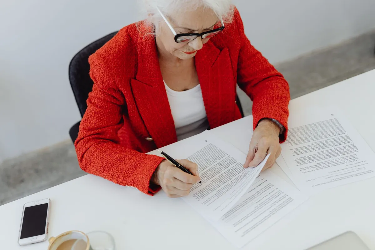 Older woman sitting at a kitchen table with paperwork and a telephone