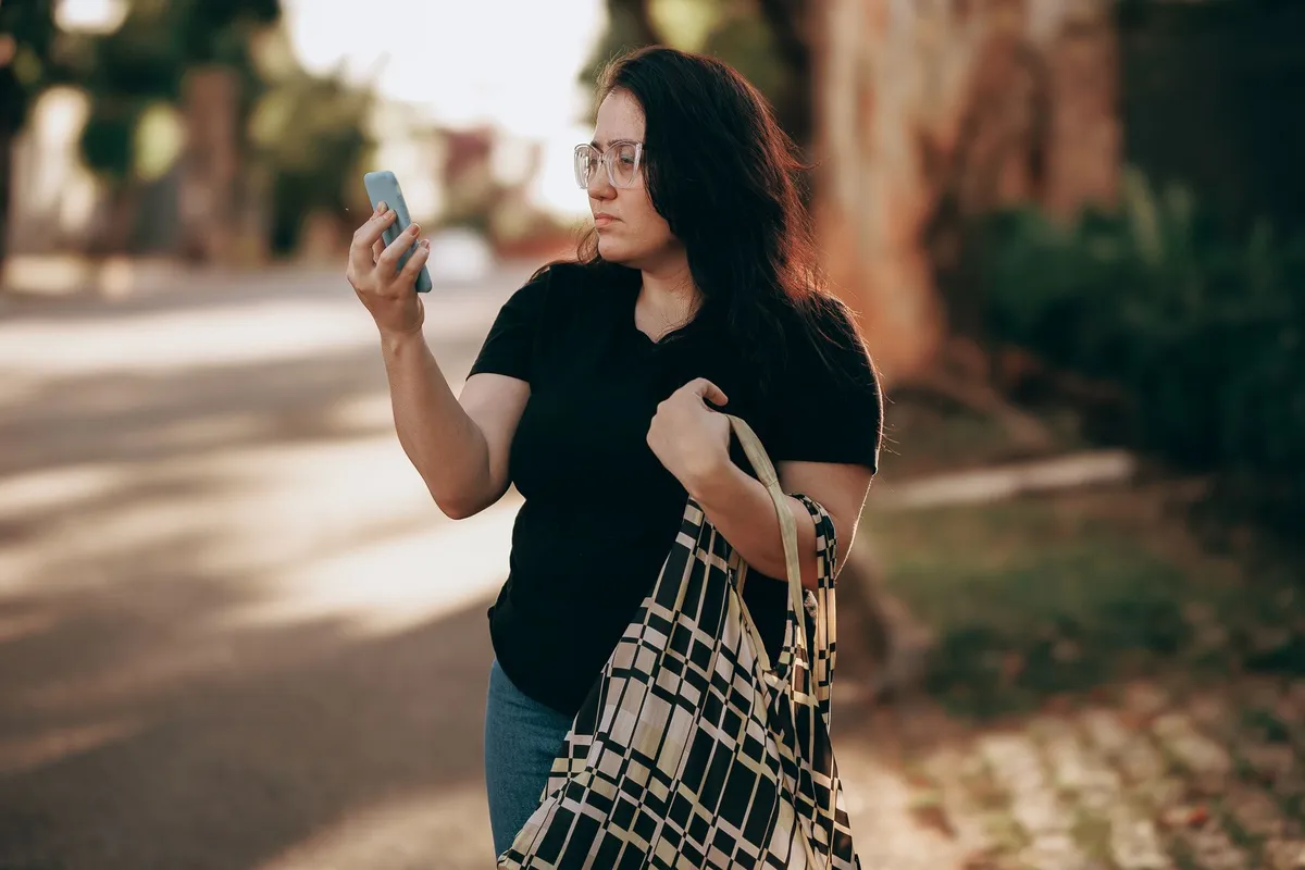 A woman with long dark hair and glasses walks outdoors while holding a checkered tote bag over her shoulder and looking at a smartphone in her raised hand