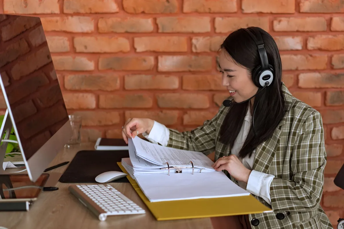 A smiling woman wearing a headset flips through papers in a binder at a desk with a computer monitor and keyboard, against a brick wall