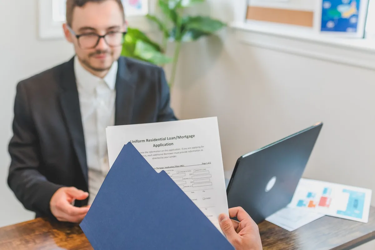 A man in a suit at a desk with a laptop looks toward a Uniform Residential Loan/Mortgage Application document being held in a blue folder by another person’s hand