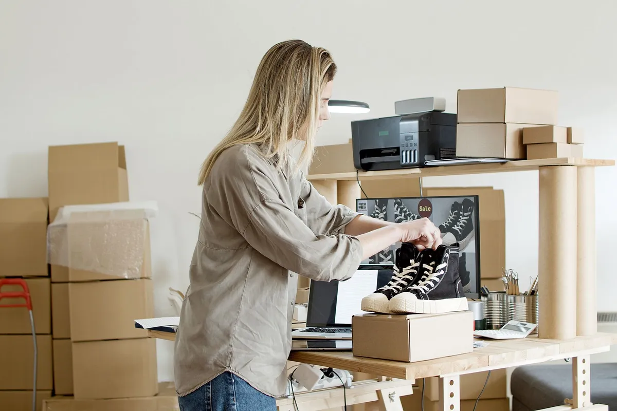 A woman ties the laces of black high-top sneakers displayed on a shipping box at a workbench surrounded by cardboard boxes, a laptop, printer, and a monitor showing shoe images with a ‘Sale’ label