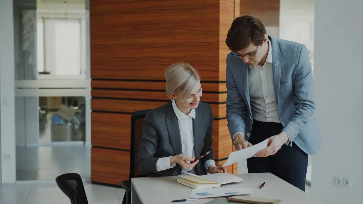 A seated woman in a gray suit and a standing man in a blazer review a document together at an office desk with notebooks and papers