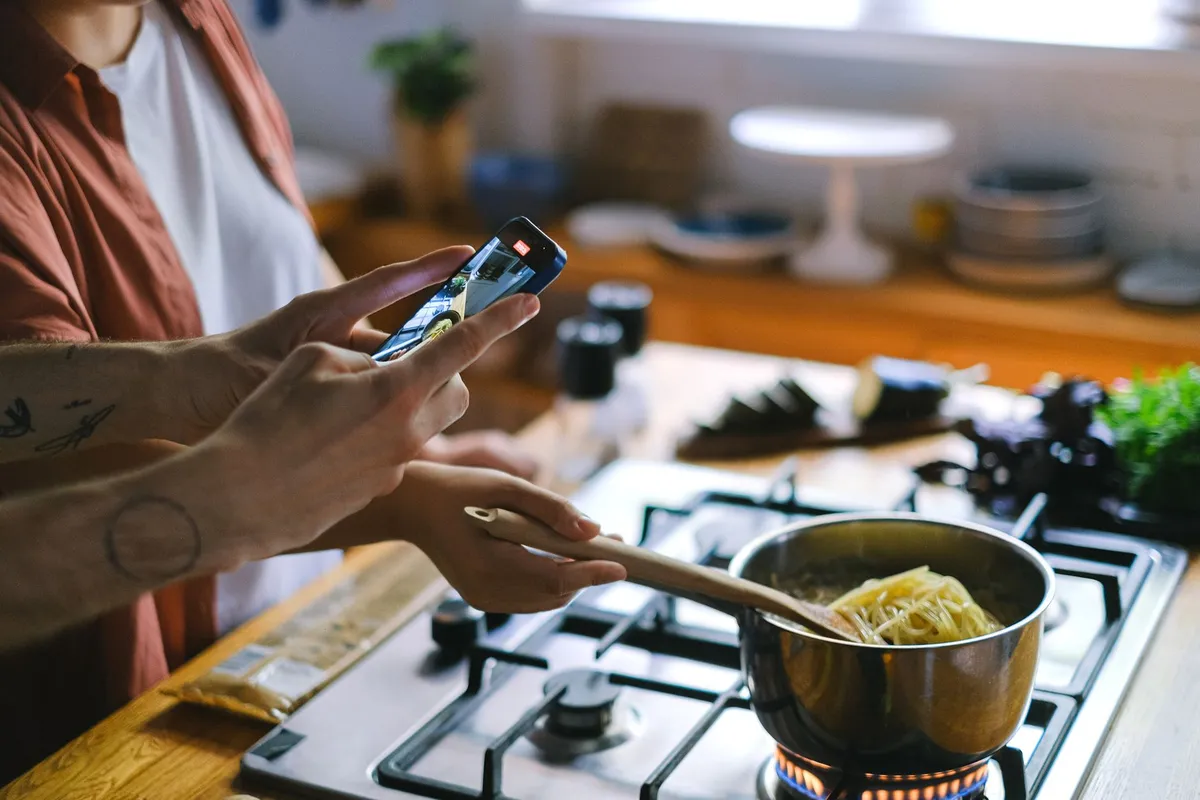 A person holds a smartphone recording video while another person stirs noodles in a pot on a gas stove