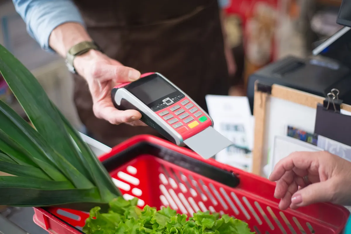 A store worker holds a card payment terminal with a card inserted over a red shopping basket filled with leafy greens at a checkout counter
