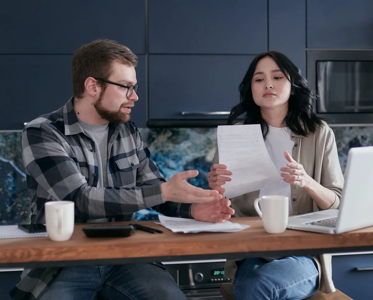 A man and woman sit at a kitchen table discussing a paper document, with coffee mugs, a phone, and an open laptop nearby